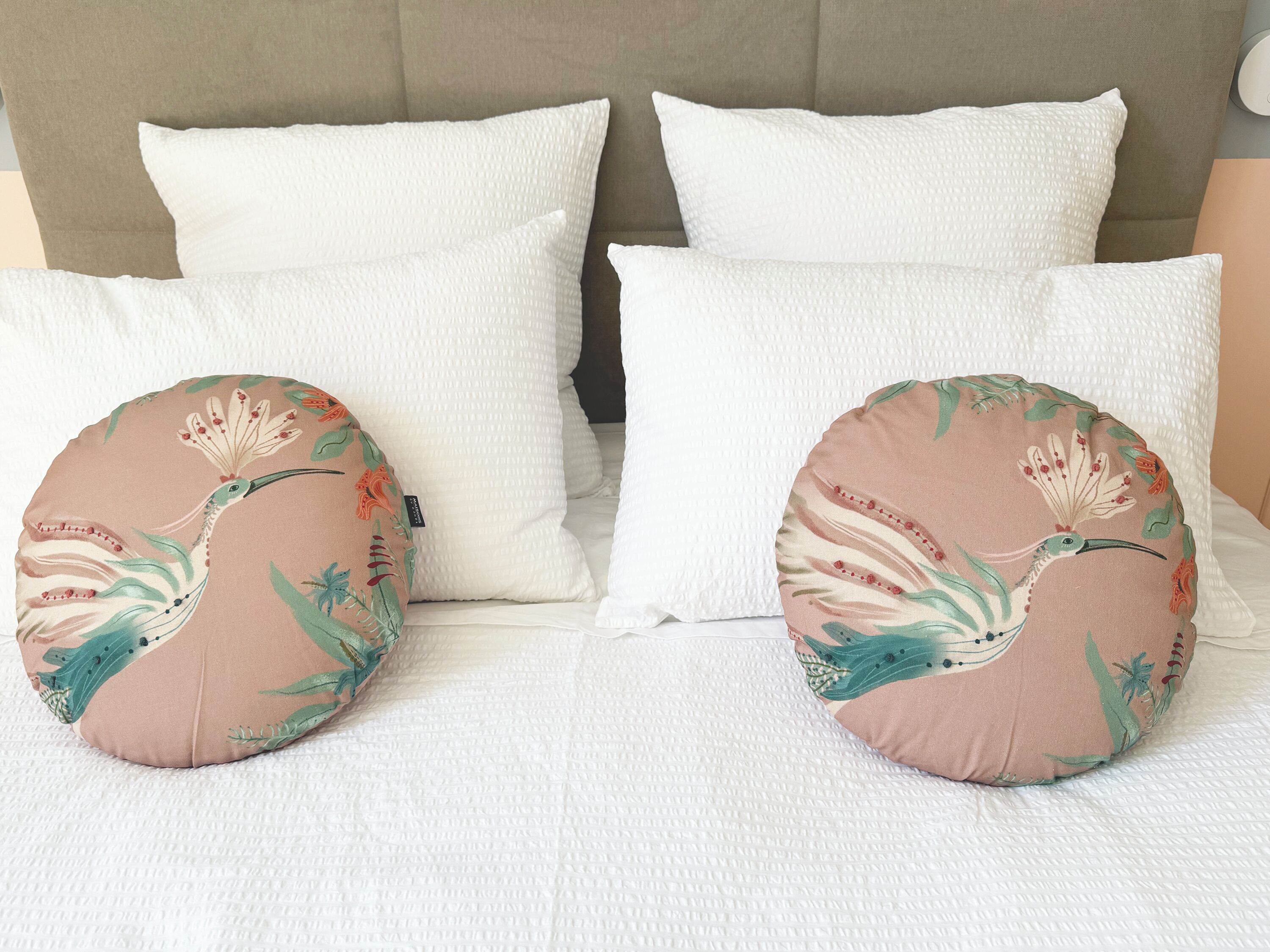 Una cama con almohadas blancas y dos cojines decorativos con motivos de pájaros y flores.