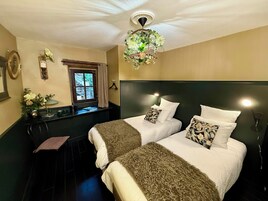 Habitación de hotel con dos camas, un candelabro, una ventana con cortinas y una pequeña mesa con una planta.
