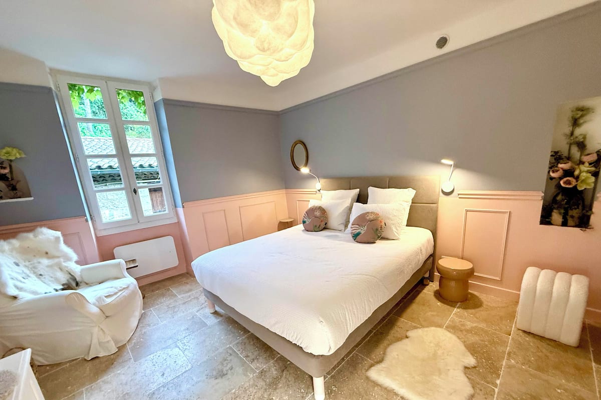 Een slaapkamer met een groot bed, een witte stoel, een klein krukje, een radiator en een raam met uitzicht op groen.