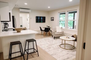 Een moderne keuken met een centraal eiland, barkrukken, een eethoek met een ronde tafel en stoelen, en een groot raam met uitzicht op groen.