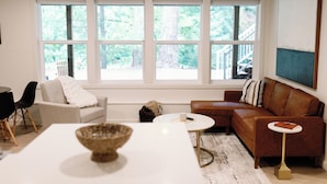 A modern living room with a white sofa, a brown leather sofa, a round coffee table, and a large window with a view of trees. - Peaceful Forest Riverfront Escape near PDX w/ Hot Tub, Kayaks, & River Access (Washougal)