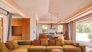 A modern living room with a large sectional sofa, wooden flooring, and a dining area with a kitchen in the background.
