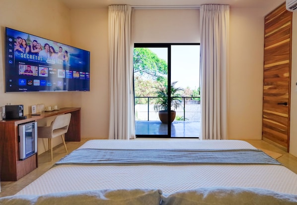 Habitación de hotel con cama, escritorio, televisor y balcón con planta.