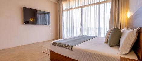 A modern hotel room with a large bed, a flat-screen TV mounted on the wall, and a large window with curtains.
