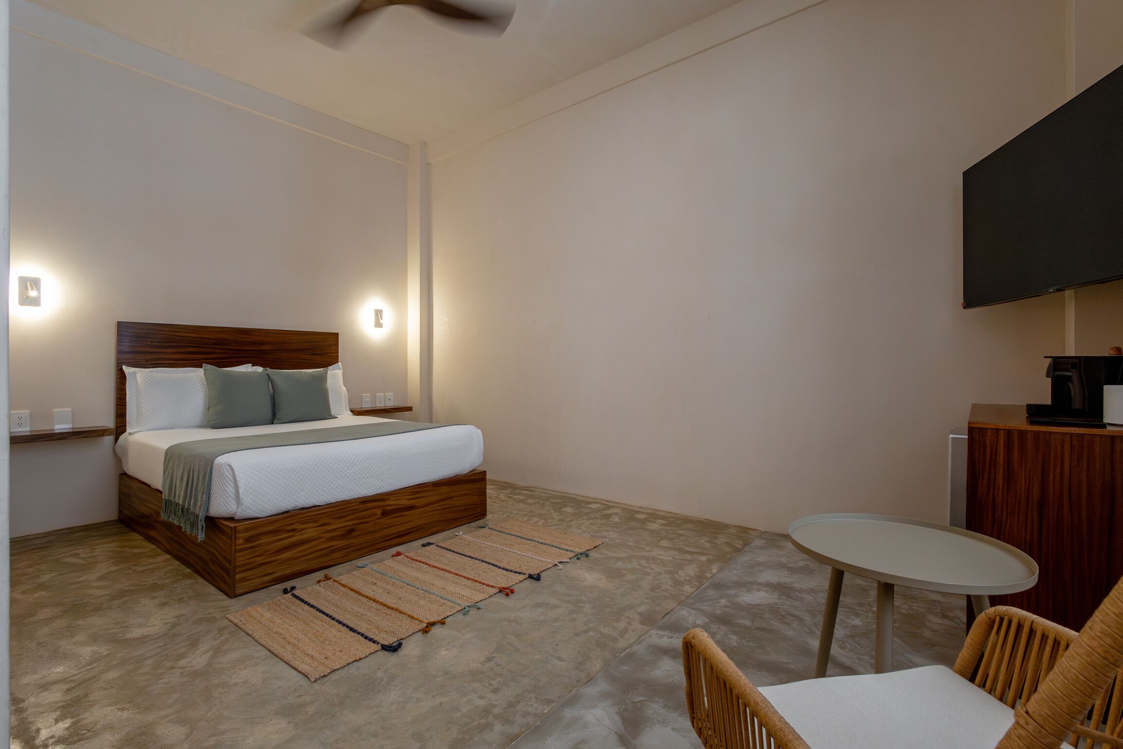 Un dormitorio moderno con una cama de madera, lámparas de noche, una mesita redonda y un televisor montado en la pared.