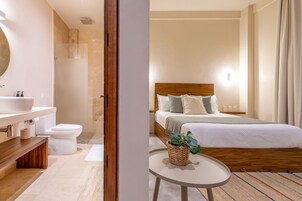 Una habitación de hotel moderna con un cabecero de madera, una mesa redonda con una planta, un espejo grande y un baño separado con inodoro y lavabo.