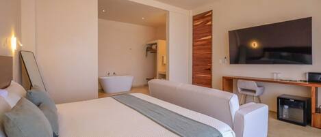 A modern hotel room with a large bed, a desk, a television, and a small refrigerator.