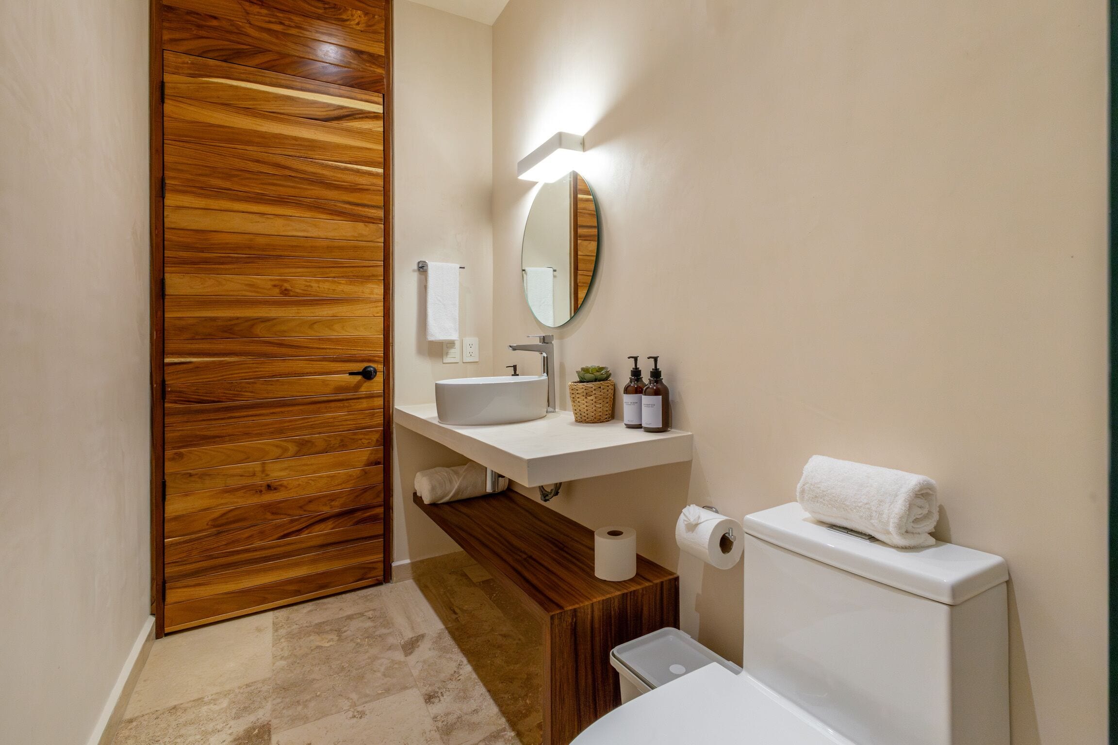 Un baño con puerta de madera, un lavamanos blanco, un espejo, un inodoro y un estante pequeño con artículos de aseo.