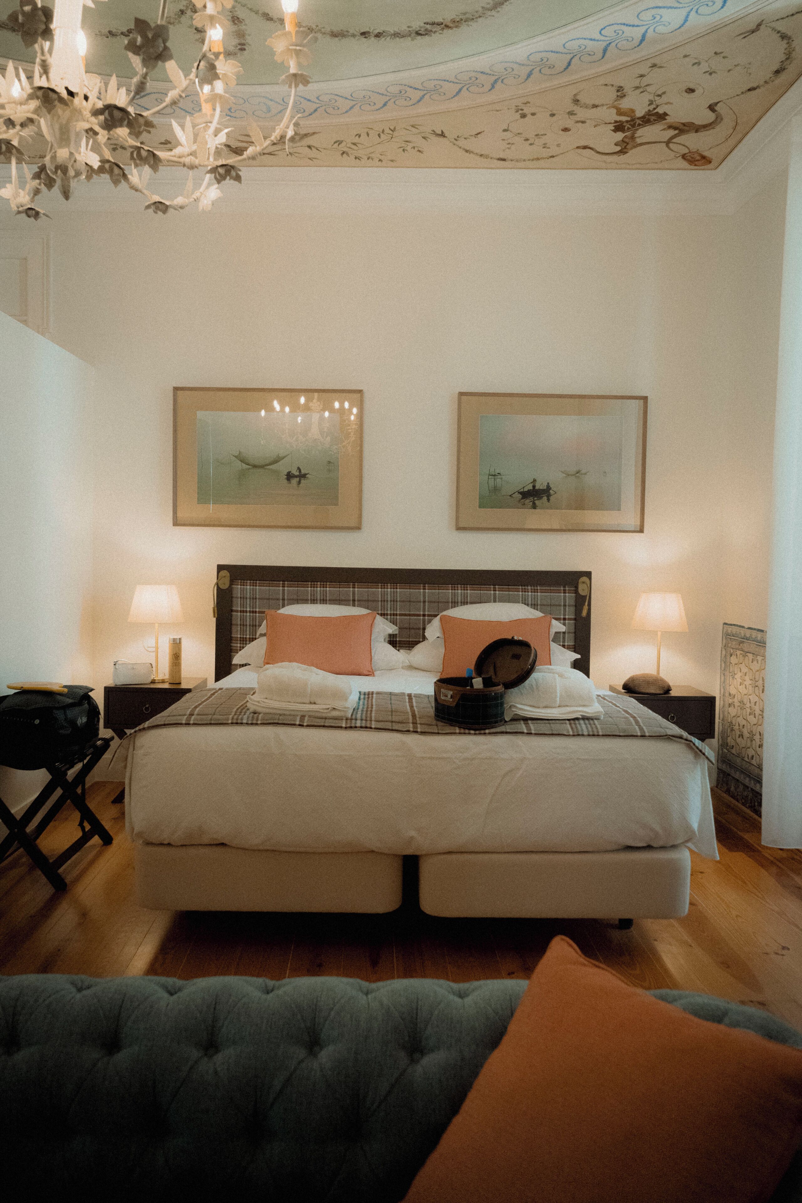 A hotel room with a large bed, two bedside tables with lamps, a chandelier, and two framed pictures on the wall.