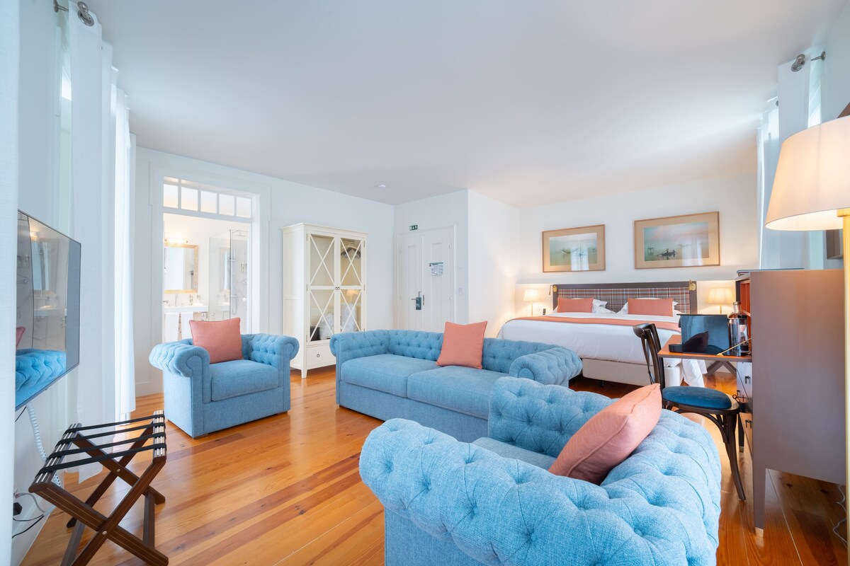 A spacious living room with a large blue sofa set, a wooden floor, and a bed in the background.