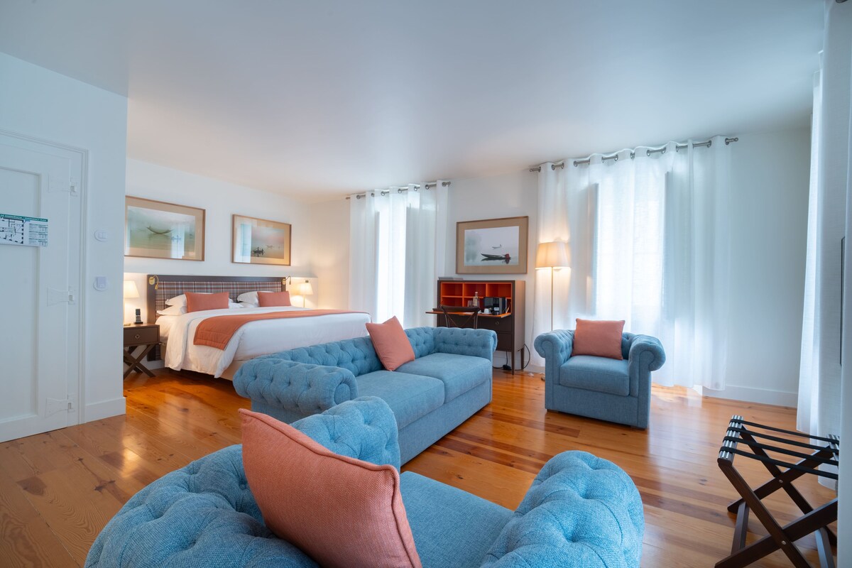 Habitación de hotel moderna con una cama grande, un sofá azul, un sillón más pequeño y piso de madera.