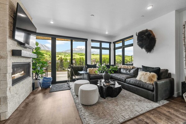 A modern living room with a fireplace, leather sofa, and large windows offering a mountain view.