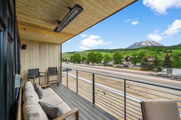 A wooden deck with a sofa and chairs, overlooking a road with vehicles and a mountain in the distance.