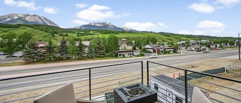 Un balcón con dos sillas y una pequeña mesa, con vistas a una calle con vehículos y edificios, y montañas al fondo.
