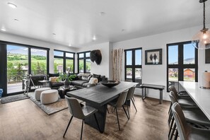 A modern living room with a large dining table, comfortable seating, and large windows offering a view of the outdoors.