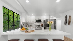 Une cuisine moderne avec des meubles blancs, un grand îlot central doté de tabourets, une vue sur la verdure grâce à de grandes fenêtres et un réfrigérateur avec distributeur d’eau.