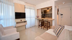 A modern living room with a flat-screen TV, a sofa, a dining area with a table and chairs, and a kitchen with a microwave and refrigerator.