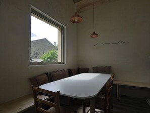 Ein Zimmer mit einem weißen Tisch, Holzstühlen, einer Bank, einem Fenster mit Blick auf Gebäude und Grünflächen sowie einer schlichten Wanddekoration.