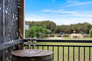 Une table en bois, avec deux verres posés sur la rambarde d’un balcon, donnant sur un espace verdoyant avec des arbres et une table de pique-nique.