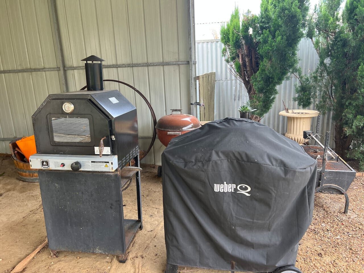 Un barbecue Weber doté d’un panneau de commande et d’une bouteille de propane protégée, installé à l’extérieur, avec un abri en métal et des arbres en arrière-plan.