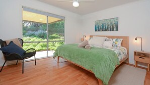 A bedroom with a large window, a bed with a green blanket, a chair, and a nightstand with a lamp.