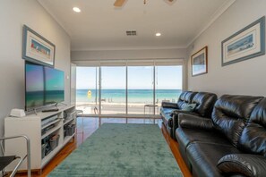A living room with a large TV, leather sofa, and a view of the beach through large glass doors.