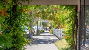 Un chemin bordé de verdure et délimité par une clôture blanche, qui mène à un bâtiment doté d’une entrée abritée.