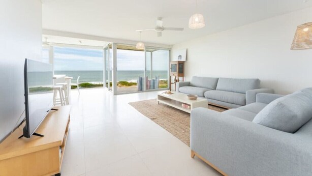 Una sala de estar moderna con un sofá grande de color gris, un televisor de pantalla plana y una mesa de centro. Hay un amplio ventanal que da a un balcón con vistas al océano.