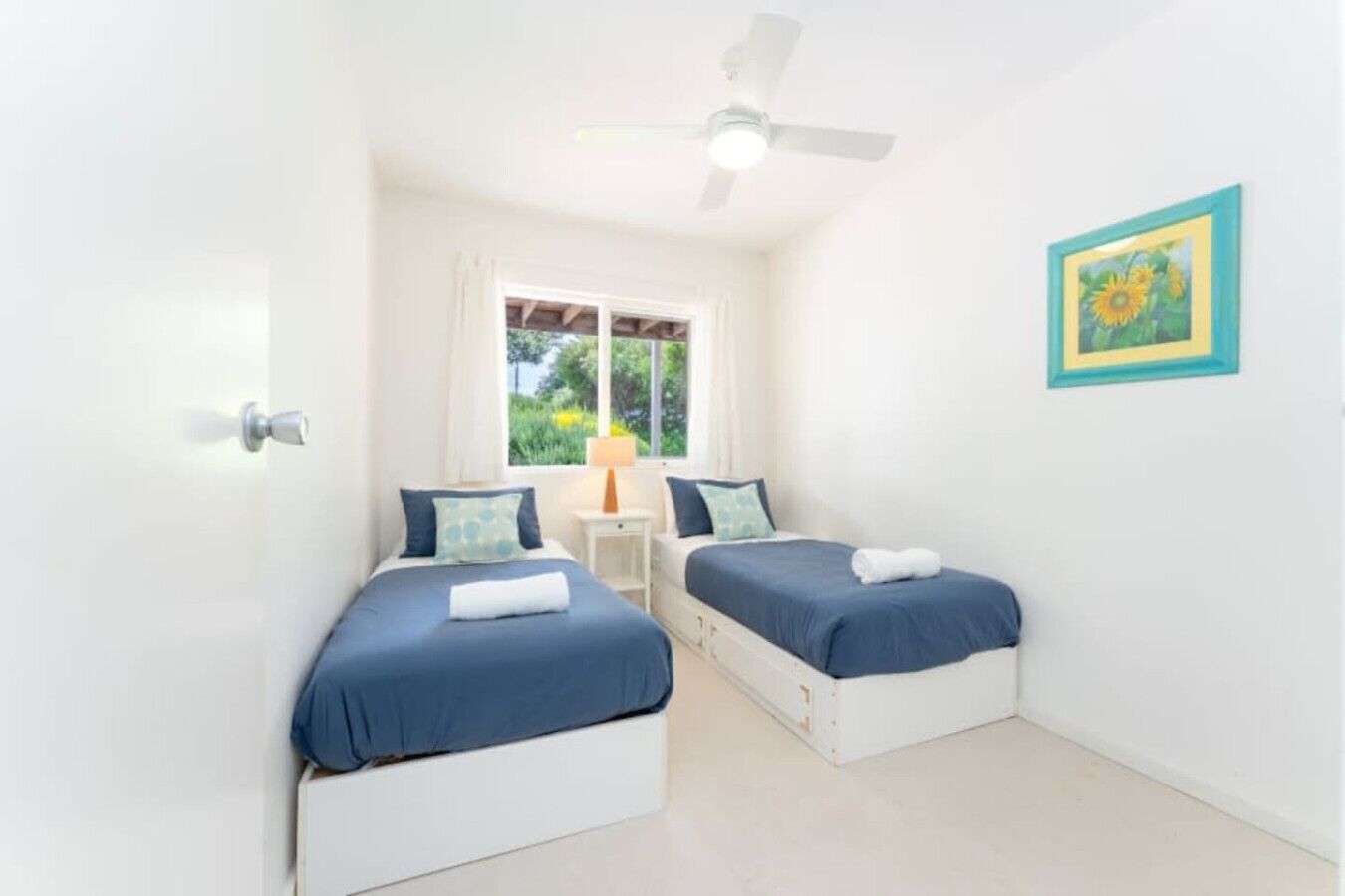 A bedroom with two beds, a ceiling fan, a window with a view of greenery, and a framed picture on the wall.