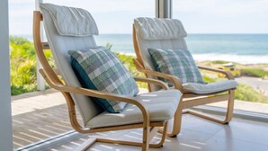 Dos sillones de madera con cojines blancos y almohadas a cuadros, ubicados en un balcón con vista al océano.