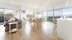 Una sala de estar moderna con un sofá, una mesa de comedor y sillas, una zona de cocina con barra y ventanas amplias que ofrecen vistas al mar.