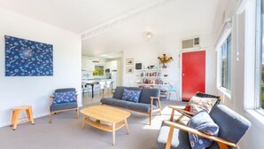 Un salon moderne avec un canapé, des fauteuils et une table basse. On aperçoit une bibliothèque remplie de livres et, au fond, une porte rouge.