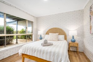 Une chambre avec une grande fenêtre, un lit doté d’une tête de lit en bois, deux tables de chevet avec des lampes et un mur en briques blanches.