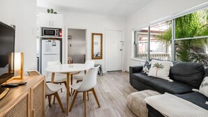 Un salon moderne avec un canapé noir, des chaises blanches, une table à manger en bois et un téléviseur posé sur un meuble.