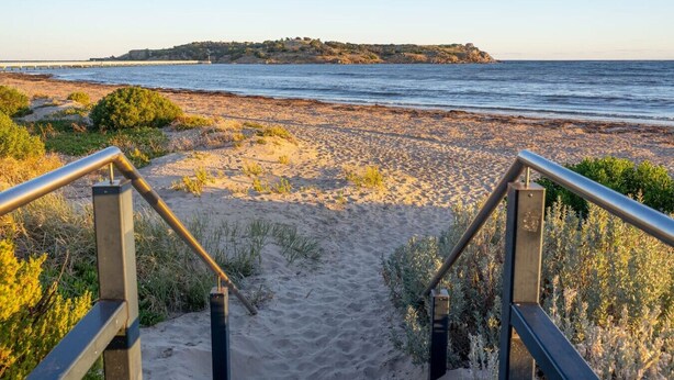 Un sentier côtier menant à une plage, avec une rambarde en métal et un terrain sablonneux.