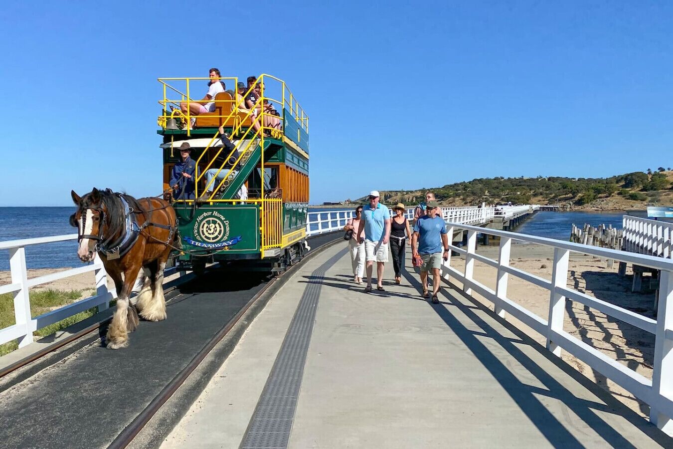 Sporvogn med passagerer trukket af en hest, langs en hvidt-indhegnet træpromenade ved havet.