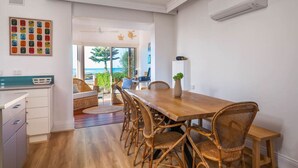 A dining area with a wooden table and wicker chairs, a white cabinet, and a view of the sea through large windows.