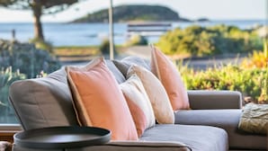 A modern living room with a grey sofa, multiple cushions, and a round table. A large window offers a view of the sea and distant hills.
