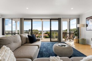 A spacious living room with a large sofa, a coffee table, and a view through glass doors to a balcony with a view of the countryside. - Hazel Beach House with ocean views (Goolwa Beach)