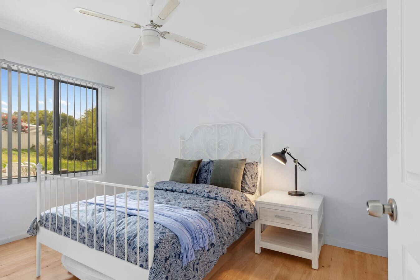 A bedroom with a bed, a nightstand, a desk lamp, a ceiling fan, and a window with a view of greenery.