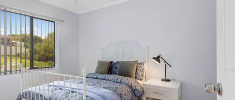 A bedroom with a bed, a nightstand, a desk lamp, a ceiling fan, and a window with a view of greenery.