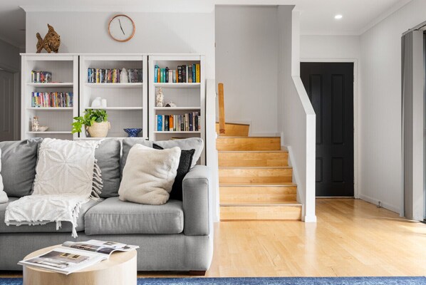 A modern living room with a grey sofa, a coffee table, and a bookshelf filled with books. A staircase leads to a dark door. - Hazel Beach House with ocean views (Goolwa Beach)