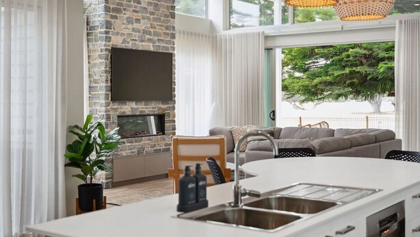 A modern kitchen with a stone wall, a built-in TV, a large island with a sink, and a view of greenery outside.