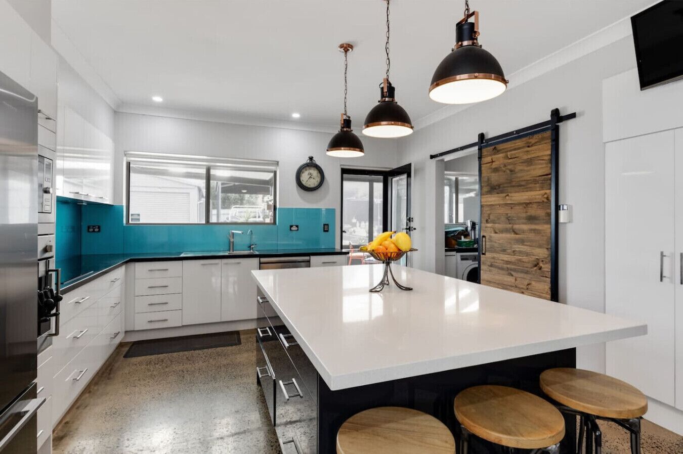 A modern kitchen with white cabinets, a large island, and a blue backsplash. There are two pendant lights, a clock, and a wooden wall panel.