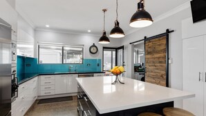 Una cocina moderna con armarios blancos, una isla grande y un salpicadero azul. Se observan dos luces colgantes, un reloj y un panel de pared de madera.