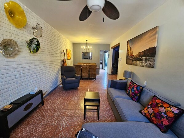 A living room with a sofa, a coffee table, and a TV. There is a brick wall with decorative plates and a ceiling fan.