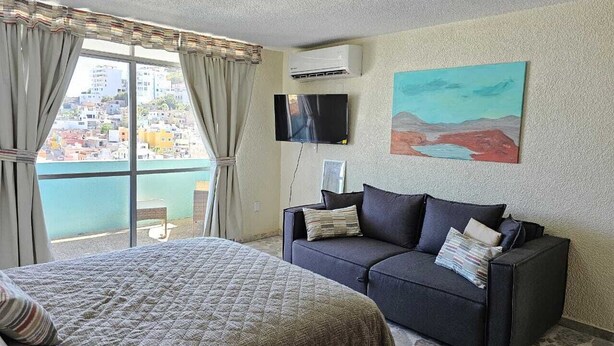 A hotel room with a bed, a sofa, a TV, and a painting on the wall. - Pool, Park & Ocean - Centro Home (Mazatlan)