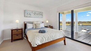 Un dormitorio con una cama grande, dos mesitas de noche, una mesita auxiliar, una ventana con vistas al agua y edificios, y una lámpara en el techo.