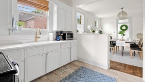 Cocina moderna con armarios blancos, microondas y cafetera sobre la encimera. Hay un comedor con mesa y sillas, y una lámpara colgante arriba.