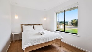 A bedroom with a wooden bed, white bedding, a small bedside table, and a large window with a view of the outdoors. - Coastal Retreat—Treetop Outlooks by Goolwa Beach (Goolwa South)